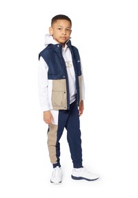 Gilet imbottito blu e beige su una felpa bianca con cappuccio, abbinato a pantaloni jogger blu e beige. Scarpe atletiche bianche completano l'outfit.