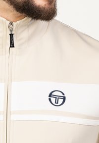 Giacca beige con zip, texture a coste, caratterizzata da strisce bianche e un logo ricamato blu scuro sul petto. Elementi in metallo della zip di colore argento visibili.