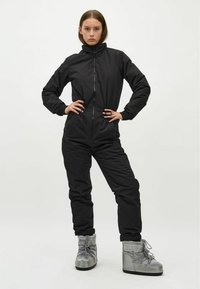 Schwarzer isolierter Overall mit hohem Kragen, Frontreißverschluss und elastischen Bündchen. Kombiniert mit grauen Schneestiefeln mit Schnürdesign und Logo.