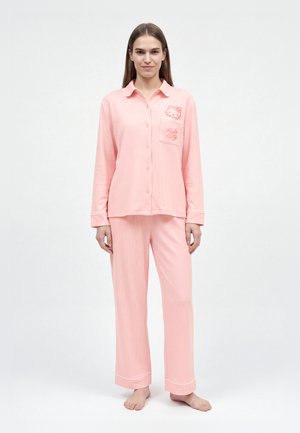 Une femme porte un pyjama Hello Kitty rose à rayures, composé d’une chemise boutonnée et d’un pantalon assorti, sur un fond blanc uni.