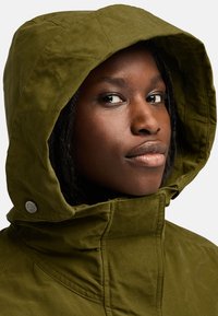 Grön huvjacka med ett vadderat huva, texturerad tyg och synlig sömnad. Har en knäppning nära kragen.