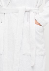 Accappatoio bianco in spugna con cintura da annodare, due tasche frontali e texture morbida. Design casual adatto per l'uso in casa.