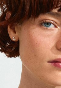 Pendiente en forma de media luna de plata con pequeñas piedras preciosas transparentes, exhibido en la oreja de un modelo sobre un fondo neutro. Se aprecia el cabello corto y oscuro.