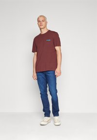 Bordo bombažna majica s plavim logotipom "wrangler", v kombinaciji z modrimi kavbojkami in belimi supergami, stoji na nevtralni podlagi.