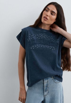 Mujer con pelo largo y oscuro que lleva una camiseta sin mangas azul oscuro con tachuelas y vaqueros de talle alto azul claro, tocándose el cuello y mirando hacia un lado.