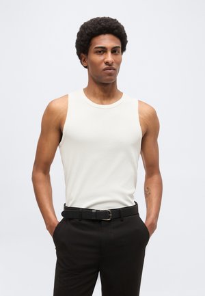 Homme avec coiffure afro portant un haut blanc sans manches et un pantalon noir, les mains dans les poches, sur un fond clair uni.