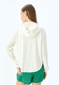 Sudadera con capucha de color beige claro, con mangas largas, corte relajado y dobladillo redondeado, combinada con unos pantalones cortos deportivos verdes. Textura de tela suave.
