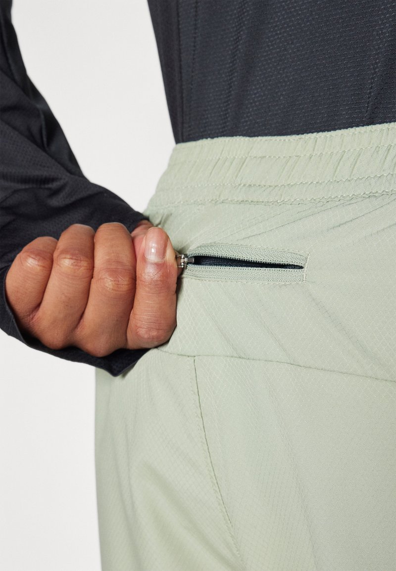 Pantalones deportivos verde claro con un bolsillo con cremallera, que presentan un patrón texturizado, cintura elástica y una parte de una camiseta negra de manga larga visible.