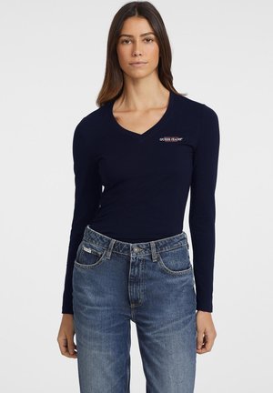 Mornarski top dugih rukava s V-izrezom u uskom kroju, s crveno-bijelim "Guess Jeans" logotipom na lijevom prsnom dijelu, u kombinaciji s plavim trapericama.