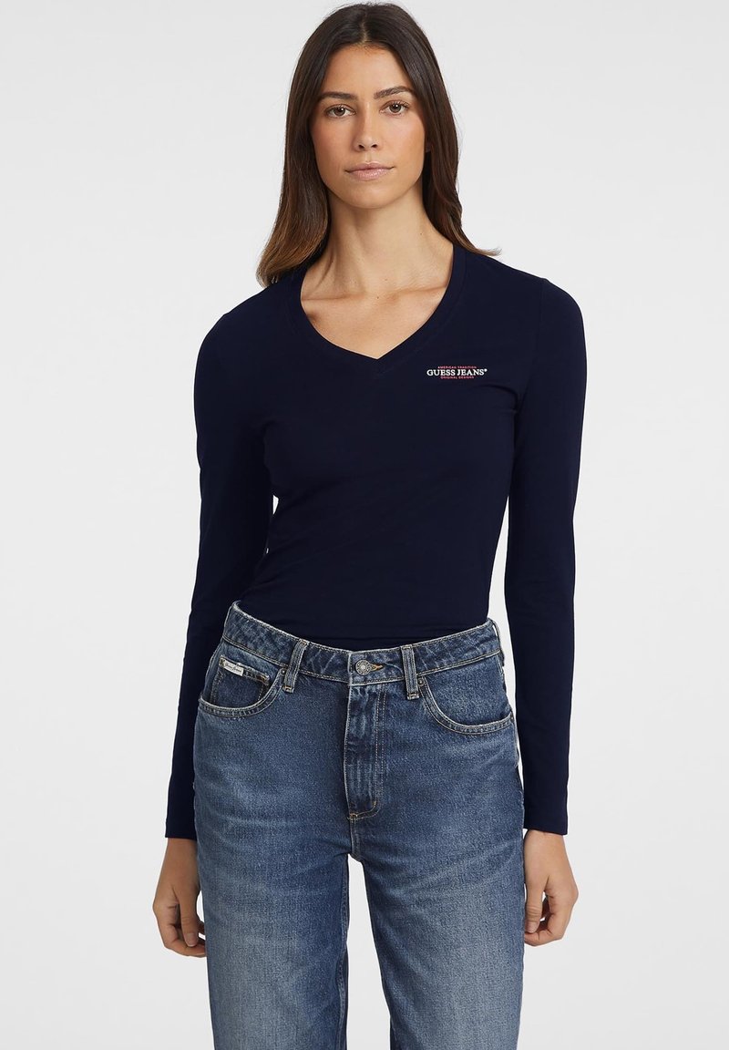 Navyblaues Langarm-V-Ausschnitt-Oberteil mit schlanker Passform, mit einem roten und weißen "Guess Jeans"-Logo auf der linken Brust, kombiniert mit blauen Denim-Jeans.