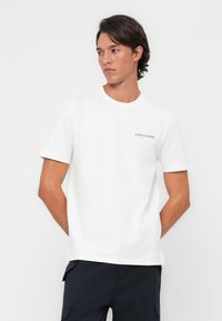 White cotton t-shirt with short sleeves, crew neckline, featuring a small black printed logo on the left chest area. Flat texture.