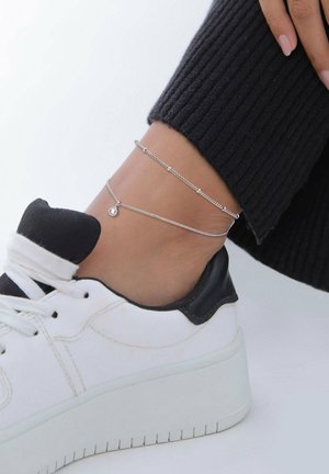 Sølv ankelarmbånd med delikat kædedesign, der har små runde accenter. Bæret over en sort ribbestrikstrømpe og en hvid sneaker.
