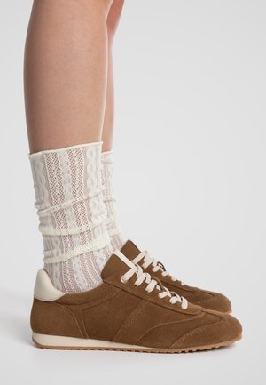 Jambes portant des chaussettes en dentelle blanches à motifs associées à des baskets en daim marron avec des lacets et semelles blanches sur un fond uni.