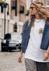 Jachetă gri din fleece cu fermoar peste un tricou alb cu o grafică cu o cutie, asortată cu pantaloni cu model. Accesoriile includ un colier și inele.