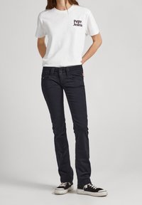 Vit t-shirt med "Pepe Jeans"-logotyp i svart, kombinerad med mörka jeans och svarta sneakers med vita sulor. Enkel, figurnära design.