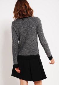 Pull gris en maille avec un motif texturé, manches longues et un ourlet côtelé noir. Porté avec une jupe noire, offrant un look superposé.