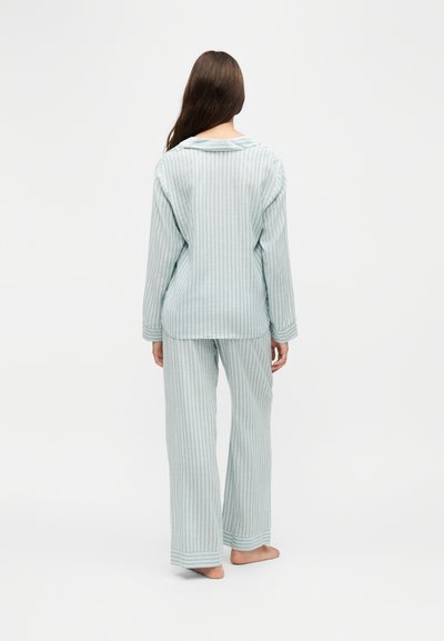 Femme debout pieds nus, de dos, portant un haut de pyjama à manches longues rayé vertical bleu clair et blanc avec un pantalon assorti.