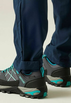 Marineblauwe broek met verstelbare boorden over grijze en turquoise waterdichte wandelschoenen, van achteren getoond tegen een effen lichte achtergrond.