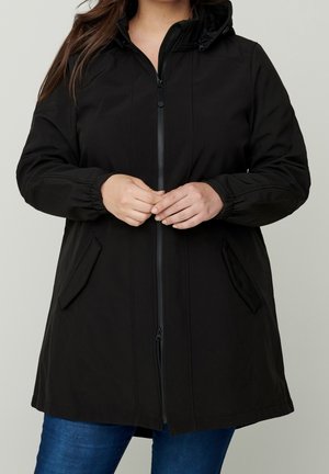 Mujer con abrigo negro con capucha y cremallera, puños elásticos y bolsillos con solapa, de pie con las manos entrelazadas sobre unos jeans azules.