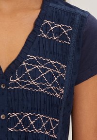 Mörkblå blus med texturerade prickar, rosa geometrisk broderi, volang vid V-ringning och knappar fram på en persons överkropp.