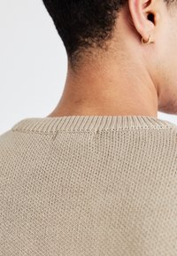 Beige sweater, der viser et struktureret strikkemønster, med en ribkant rundt om halsen. En guldringeørering er synlig i øret.