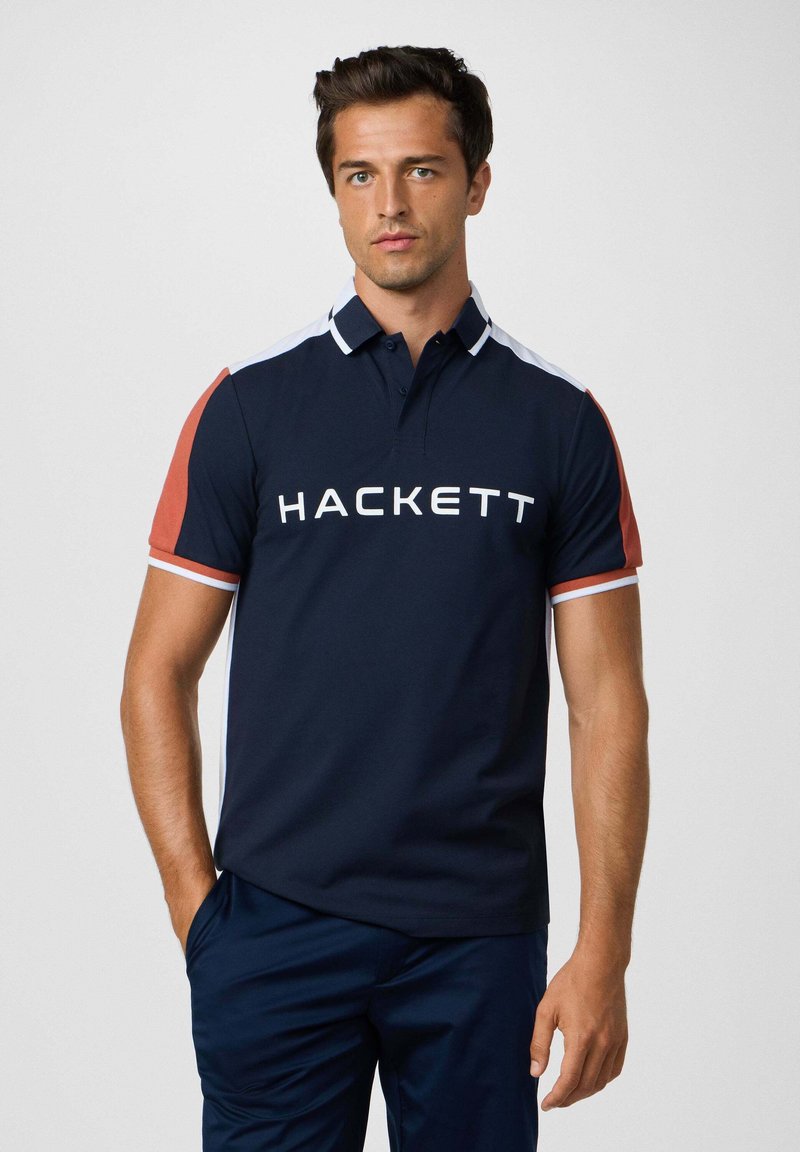 Marineblauwe polo met rode en witte accenten, met een groot wit "HACKETT" logo op de borst. Gemaakt van gladde katoenstof.