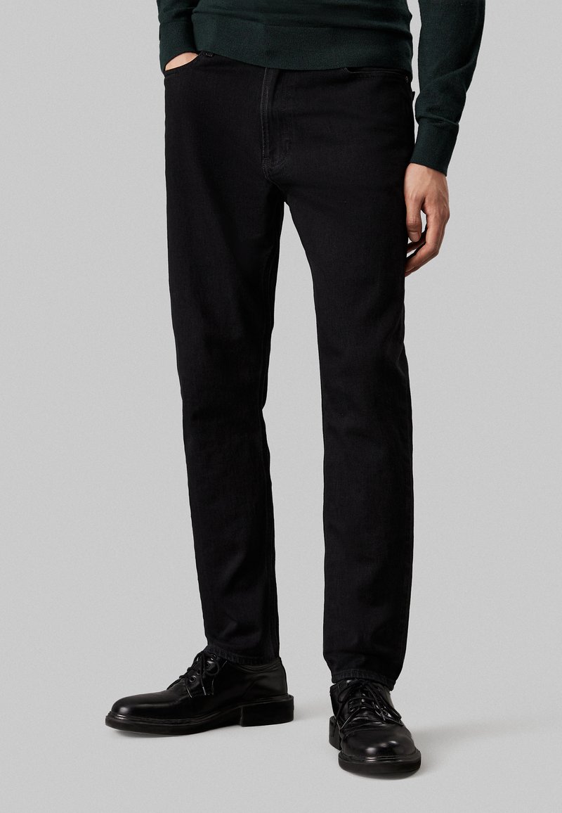 Pantaloni in denim nero con una vestibilità slim, dotati di tasche anteriori e design classico. Indossati con scarpe nere per un look coerente.