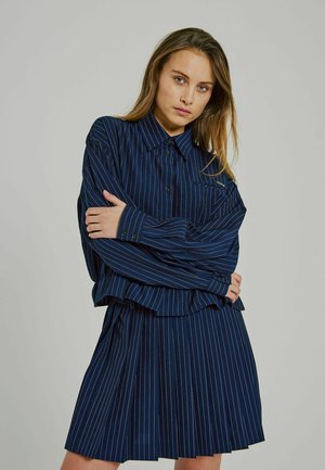 Navy blue, pinstriped shirt with a collared neckline and button closure, paired with a pleated skirt. Fabric appears smooth and light.
