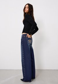 Hoch taillierte, weit geschnittene Jeans aus dunkelblauem Denim. Verziert mit dekorativen Nieten entlang der Seite und einem Sternmuster auf der Gesäßtasche.