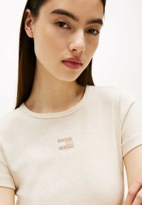 Beżowa, prążkowana bluzka z krótkim rękawem z małą prostokątną naszywką z napisem "TOMMY JEANS" w białym i brązowym kolorze.