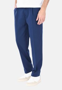 Pantaloni blu navy con una vestibilità rilassata, dotati di due pieghe frontali, tasche laterali e un tessuto morbido; indossati con sneaker bianche.