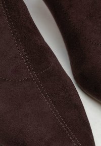 Brązowe zamszowe buty na wysokim obcasie z ostrym noskiem. Charakteryzują się zakrzywionym przeszyciem i gładką fakturą. Zbliżenie na szczegóły buta.