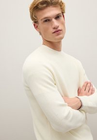 Cremefarbener Strickpullover mit gerippter Struktur, langen Ärmeln und Rundhalsausschnitt. Verfügt über kontrastierende graue Akzente an den Bündchen.