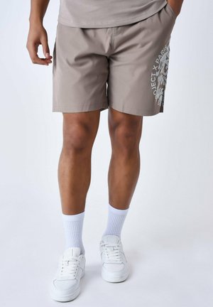 Bañador beige con cintura elástica, bolsillos laterales y un estampado gráfico en la parte inferior de la pierna derecha. Combinado con zapatillas blancas y calcetines.
