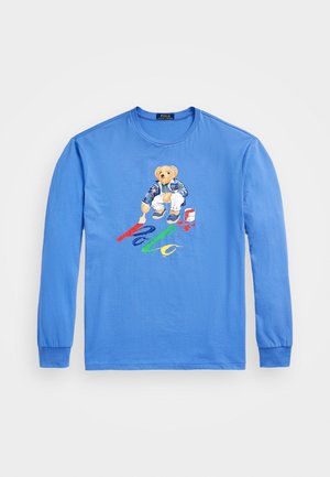 Chemise bleue à manches longues avec un ours peignant le mot "Polo" en rouge, bleu, vert et jaune sur le devant.