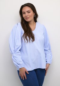 Jasnoniebieska bluzka w paski z dekoltem w kształcie litery V oraz marszczonymi, bufkami. Materiał wydaje się gładki i lekki. Połączona z ciemnymi dżinsami.