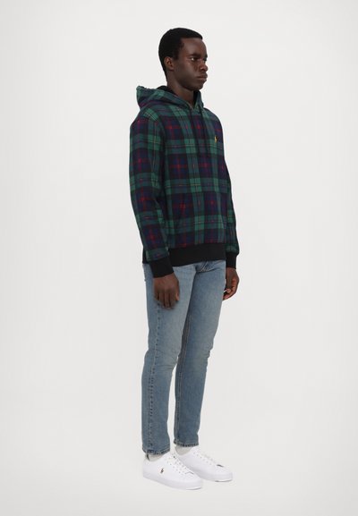 Uomo in piedi che indossa una felpa a quadri verde e blu, jeans azzurri chiari e sneaker bianche su uno sfondo bianco semplice.
