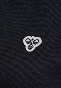 Sort stof med en lille, hvid broderet bi-logo, der har sorte detaljer på vinger og ansigt. Teksturen er glat og tæt vævet.