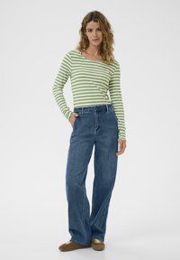 Zeleno in belo prugasta dolga majica s robom izrezom, v kombinaciji z modrimi širokimi hlačami in rjavimi čevlji, nevtralna ozadja.