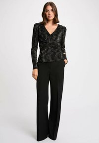 Top nero a maniche lunghe con scollatura a V, caratterizzato da un design anteriore annodato. Il tessuto ha un motivo lucido e testurizzato. Abbinato a pantaloni neri a gamba larga.