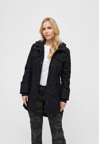 Svart parkajakke med hette, med frontlommer, snor i midjen og lange ermer, bæres over en hvit skjorte og kamuflasjebukser.