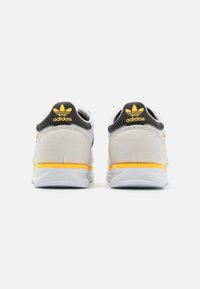 Baskets Adidas avec un dessus gris et blanc, des accents jaunes, et une languette à l'arrière texturée avec un logo. Semelle en caoutchouc pour une meilleure accroche.