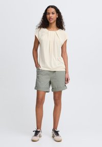 Blouse plissée crème clair, associée à un short olive roulé. Accompagnée de baskets à lacets beiges et noires.