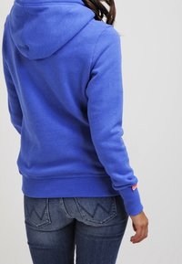 Sudadera azul con un acabado texturizado, que presenta una capucha con cordón y puños acanalados. Combinada con unos jeans que muestran costuras en zigzag.