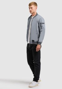 Homme portant une veste bombardier gris clair, un T-shirt blanc, un pantalon noir et des baskets blanches, debout devant un fond uni.