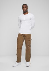 Chemise henley blanche à manches longues avec patte de boutonnage, associée à un pantalon cargo marron avec plusieurs poches, et des baskets blanches.
