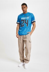 Blå sporty t-skjorte med "WROCLAW" og nummer "24" i svart, kombinert med beige cargo-bukser og hvite sneakers.