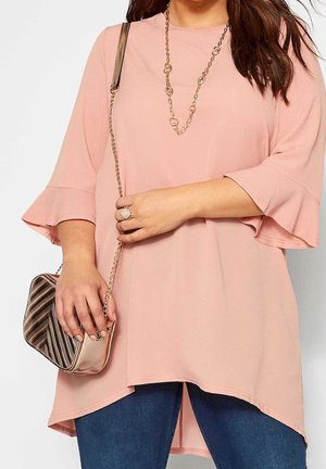 Túnica rosa claro con mangas acampanadas, tela texturizada y dobladillo asimétrico; combinada con un bolso de hombro metálico a rayas.