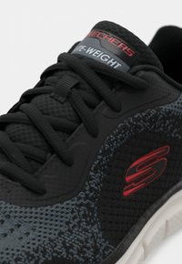 Lightweight black athletic shoe with textured fabric, reinforced sides, and a grey and black patterned design. Features red Skechers logo.