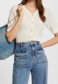 Biały wełniany kardigan z krótkim rękawem i falbaniastym dekoltem, zapinany na złote guziki, w połączeniu z dżinsami niebieskimi o wysokiej talii oraz niebieską pikowaną torebką.